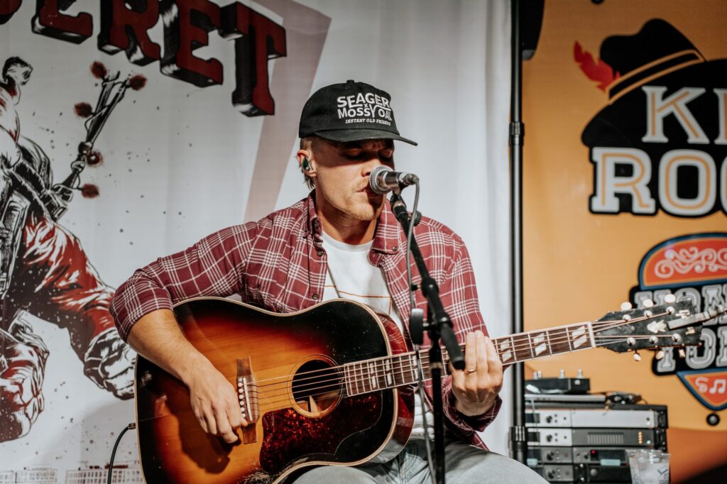Musician singing and playing an acoustic guitar on stage in front of the Rock the Green Beret backdrop