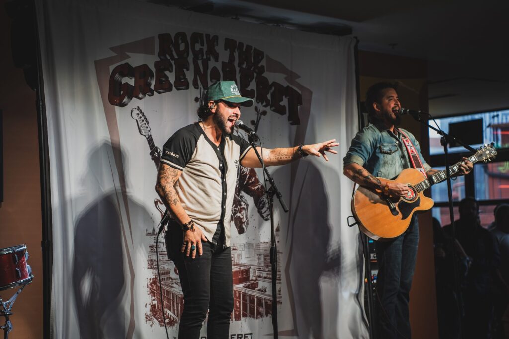 Two musicians performing on stage in front of the Rock the Green Beret banner